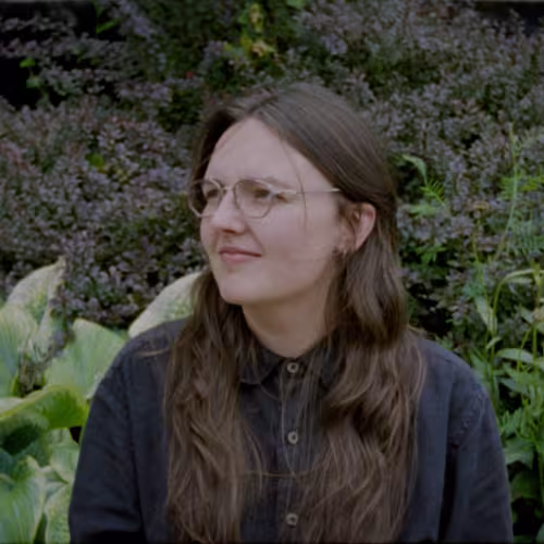 Brielle is sat in front of a garden looking to their left. They have long straight brown hair, wire glasses, and a black button down on.