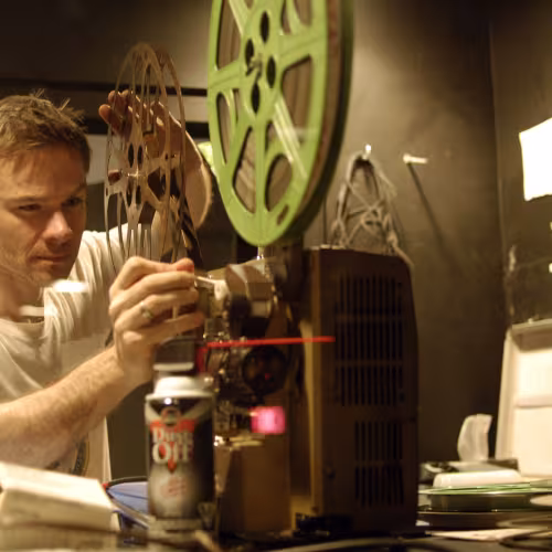 Chris is behind a 16mm projector tuning it up for projection. He has short brown hair and a white t-shirt on.